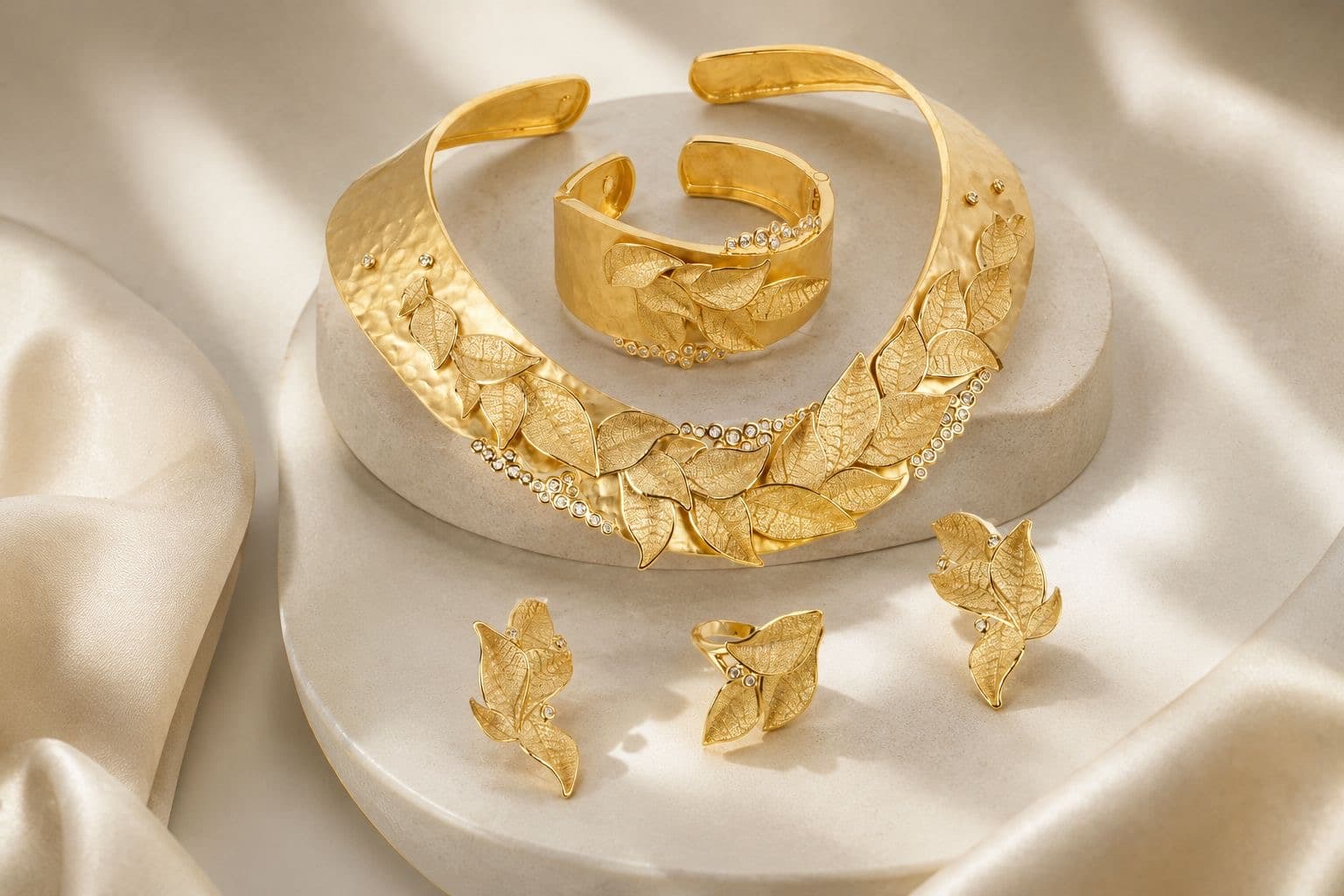 A matte gold leaf-motif suite — collar necklace, cuff, ring and earrings — arranged on ivory stone